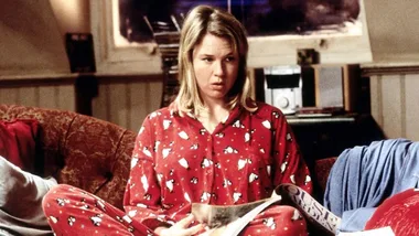 A woman in red pajamas with white patterns sits on a couch, holding a magazine, looking distraught.