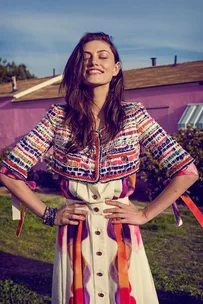 Smiling woman in colorful jacket and dress stands outside, hands on hips, against a purple house backdrop.