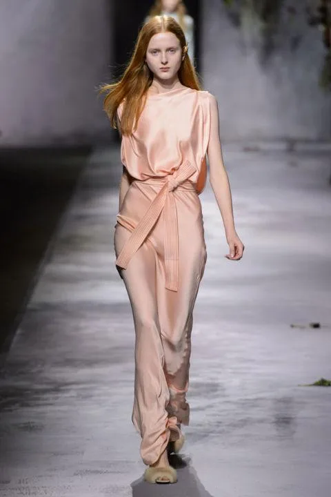 A model walks down a runway in a flowing peach gown, with long red hair cascading over shoulders in a minimalist setting.