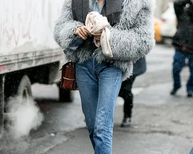 A person wearing a fluffy gray jacket, scarf, and jeans on a city street.