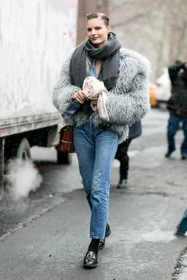 A person wearing a fluffy gray jacket, scarf, and jeans on a city street.