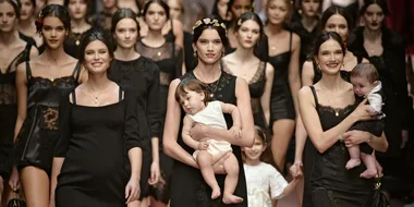 Models in black outfits walk the runway at a Dolce & Gabbana fashion show, with two models carrying babies.