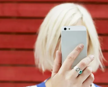 Person with platinum blonde hair taking a selfie with a smartphone against a red wooden background.