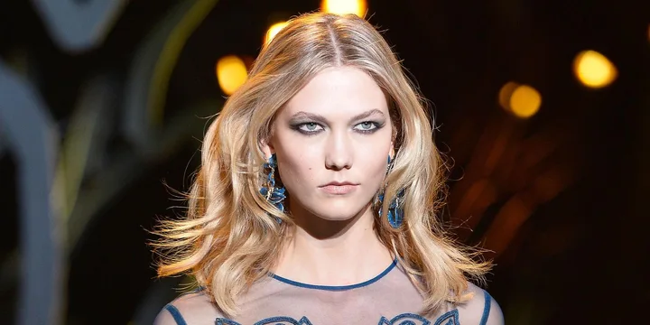 Blonde woman in a blue dress with blue earrings on a runway with blurred lights in the background.