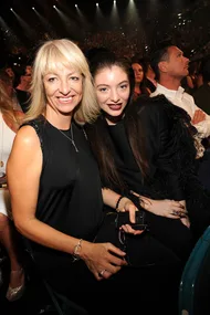 Two women sitting closely at a crowded event, both smiling at the camera. One with blonde hair, the other with dark hair.