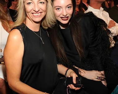 Two women sitting closely at a crowded event, both smiling at the camera. One with blonde hair, the other with dark hair.