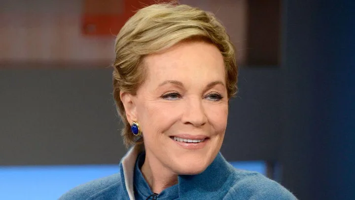 Julie Andrews smiling, wearing blue top and earrings, indoors with a blurred background.