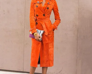 Jourdan Dunn in an orange trench coat, holding a clutch and wearing black heels, standing against a beige background.