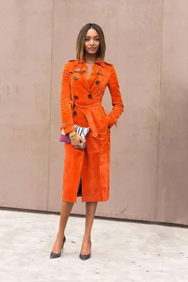 Jourdan Dunn in an orange trench coat, holding a clutch and wearing black heels, standing against a beige background.