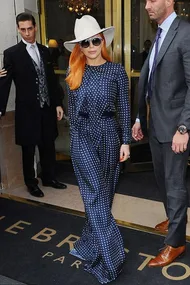 A person in a blue polka dot outfit, orange hair, white hat, and sunglasses, exits a building with security personnel.