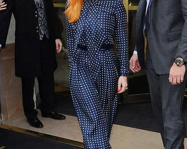 A person in a blue polka dot outfit, orange hair, white hat, and sunglasses, exits a building with security personnel.