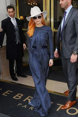 A person in a blue polka dot outfit, orange hair, white hat, and sunglasses, exits a building with security personnel.