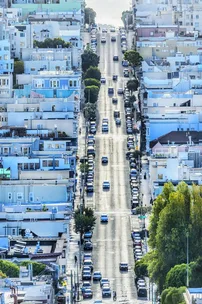 Street view of a hilly, residential neighborhood in San Francisco, lined with cars and pastel-colored buildings.