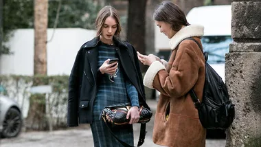 Two women in winter coats standing outdoors close together, looking at their smartphones.