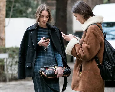 Two women in winter coats standing outdoors close together, looking at their smartphones.