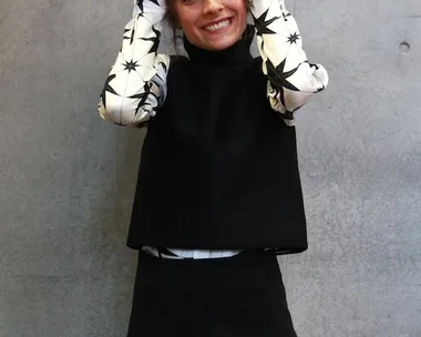 A woman wearing a black and white outfit makes peace signs with both hands against a concrete wall background.