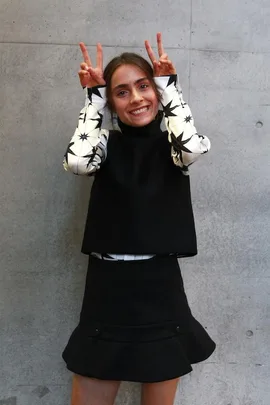 A woman wearing a black and white outfit makes peace signs with both hands against a concrete wall background.