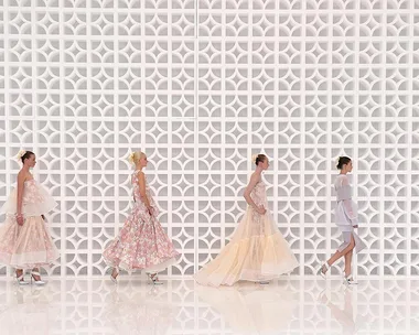 Models in pastel dresses with floral prints walking down a runway against a white geometric patterned wall.