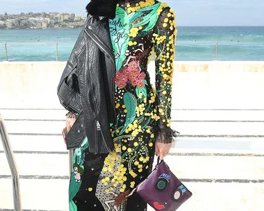 Woman in vibrant floral dress and sunglasses holds quirky face-patterned clutch, with ocean backdrop.