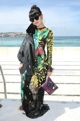 Woman in vibrant floral dress and sunglasses holds quirky face-patterned clutch, with ocean backdrop.