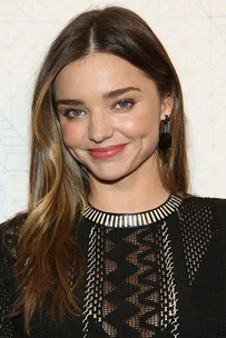 A woman with long hair smiles, wearing a black dress with intricate designs and statement earrings against a patterned backdrop.