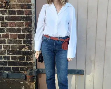 Woman in white blouse, blue jeans with red sash, and black heels standing against a brick wall and beige door.