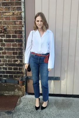 Woman in white blouse, blue jeans with red sash, and black heels standing against a brick wall and beige door.