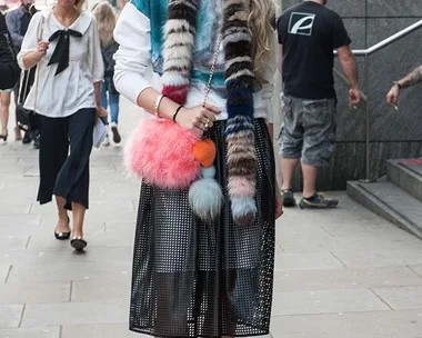 Street fashion: Woman with sunglasses, colorful fur scarf, and a pink pom-pom bag on a city sidewalk.