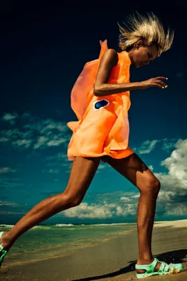 A person in a vibrant orange dress runs energetically on a sunny beach with a blue sky background.