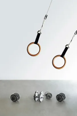 Gymnastics rings hanging above a set of four dumbbells on a gray surface.