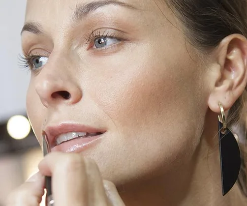 Woman applying makeup with brush, wearing black earrings, focused expression.