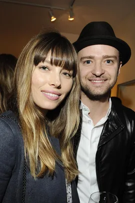 A smiling couple poses together, the woman with long hair and bangs, and the man wearing a black hat and a leather jacket.