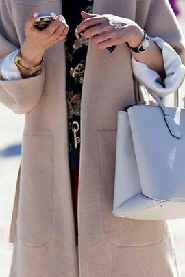 Person holding a smartphone and a white handbag, wearing a beige coat.