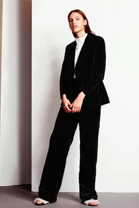 A woman wearing a black suit with a white top stands against a white and beige background.
