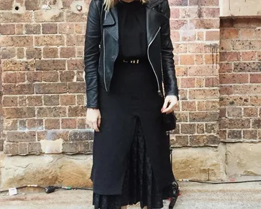 Woman in black leather jacket and black dress stands in front of a brick wall, looking down, streetstyle fashion.