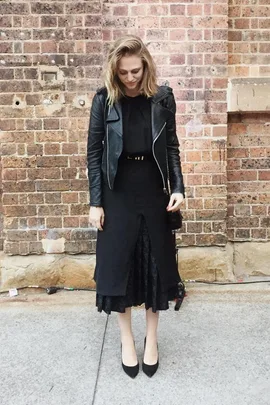 Woman in black leather jacket and black dress stands in front of a brick wall, looking down, streetstyle fashion.