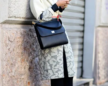 Woman in stylish coat and sunglasses leaning against wall, checking phone; holding black handbag.