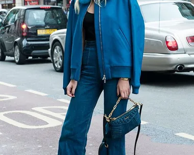 Woman in blue jacket and jeans with handbag, standing on urban street with cars and buildings in background.