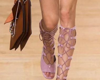 A woman wearing light pink lace-up sandals walks on a runway, holding a brown handbag.