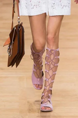 A woman wearing light pink lace-up sandals walks on a runway, holding a brown handbag.