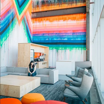 A modern office lounge with colorful geometric wall art, gray seating, and a person sitting on a couch.