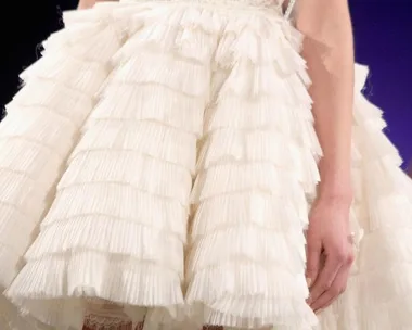 Close-up of a short, tiered ruffled white bridal dress with a textured pattern, worn by a model on a runway.