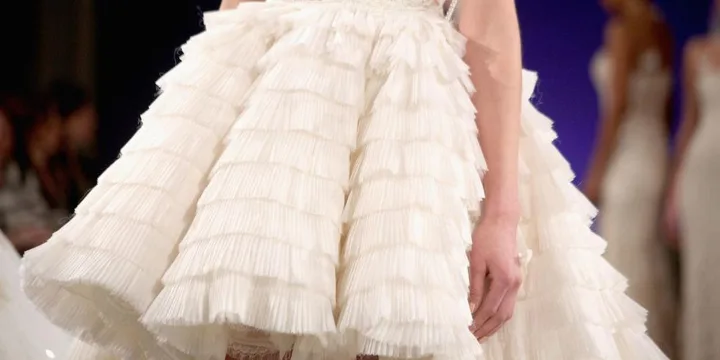 Close-up of a short, tiered ruffled white bridal dress with a textured pattern, worn by a model on a runway.