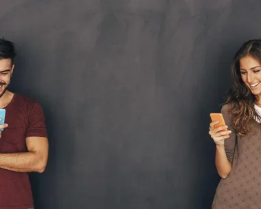 A man and a woman standing and smiling at their smartphones in front of a dark gray wall.