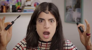 A woman with wide eyes and open mouth holding mascara wands with a blotch of makeup on her forehead, wearing a striped shirt.