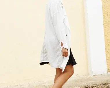 A woman in a white coat and black boots walks on a paved path with a yellow wall background, smiling and wearing sunglasses.