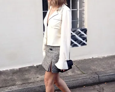 Person wearing a white jacket, grey skirt, black heels, and carrying a black bag walking on a street near a building.