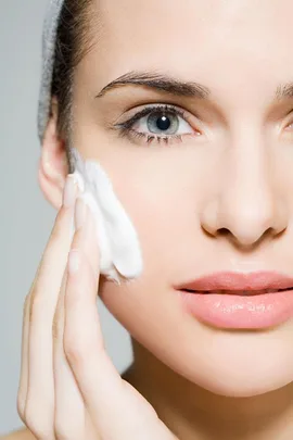 A woman applying a foamy white cream to her face, focusing on her clear complexion and skincare routine.