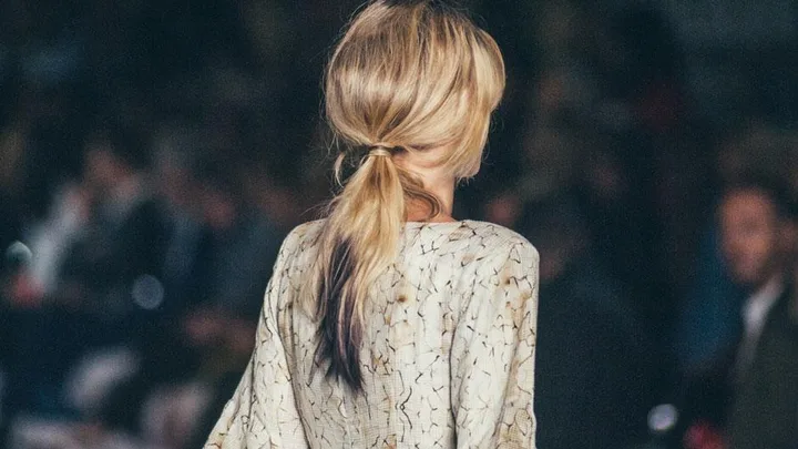 A model with blonde hair in a voluminous low-slung ponytail on a runway, wearing a light patterned outfit.