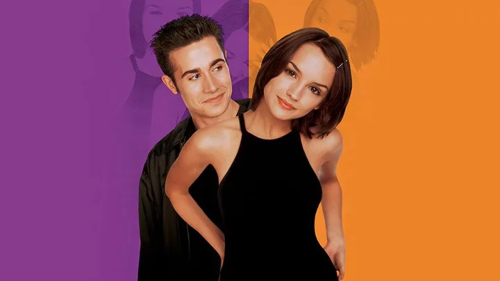 A young man and woman from "She's All That" stand close together with a colorful background.